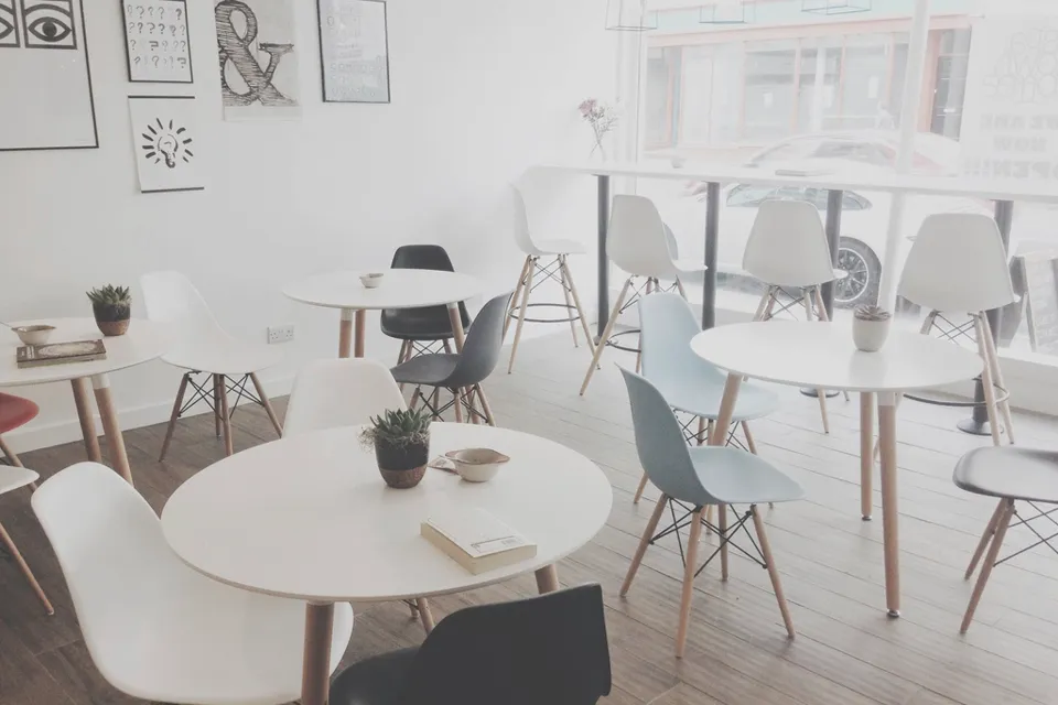 Bright minimalist white cafe with round white tables, white, grey and blue chairs on wooden legs, small succulents and posters by a large front window.
