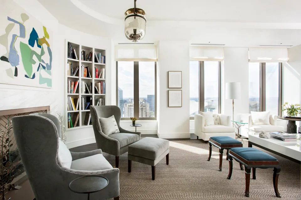 Bright high-rise apartment living room with grey wingback armchairs, built-in bookshelves, abstract art, blue stools and city views through tall windows