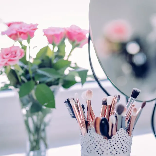 Rose gold makeup brushes in a white lace-pattern holder on a vanity beside a round mirror and pink roses